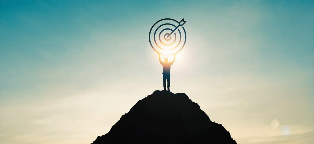 Person standing on hilltop holding an illustration of a target with arrow in the bullseye above their head with the sun shining behind them